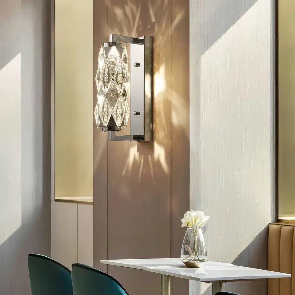 A wall-mounted crystal sconce illuminates a modern dining area with a geometric light pattern. A small table with two green chairs is adorned with a clear vase holding white flowers. Minimalistic decor creates a serene ambiance.