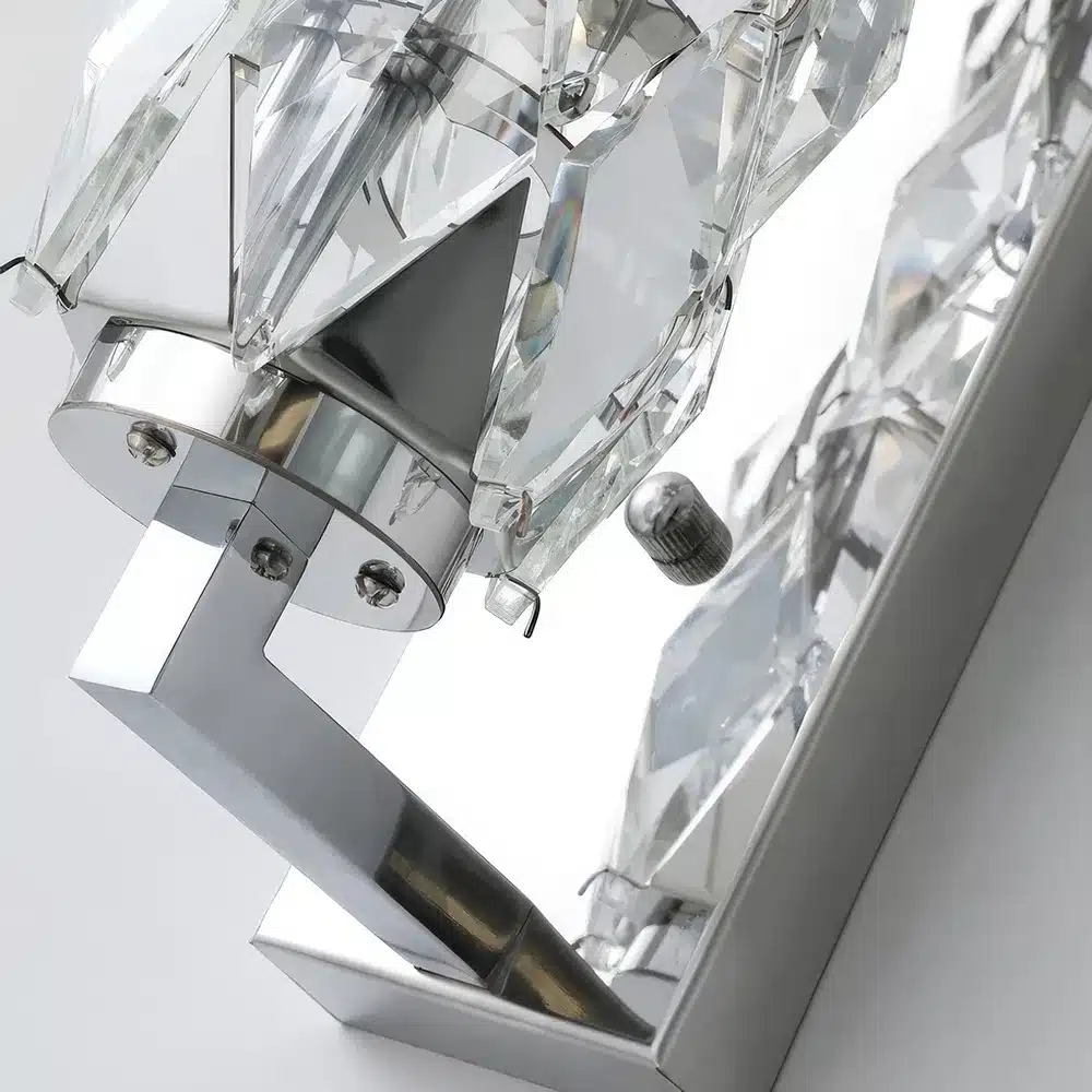 Close-up view of a modern, geometrically-shaped wall sconce with clear crystal and chrome detailing, reflecting light. The angular design and metallic finish give it a sleek, contemporary appearance.