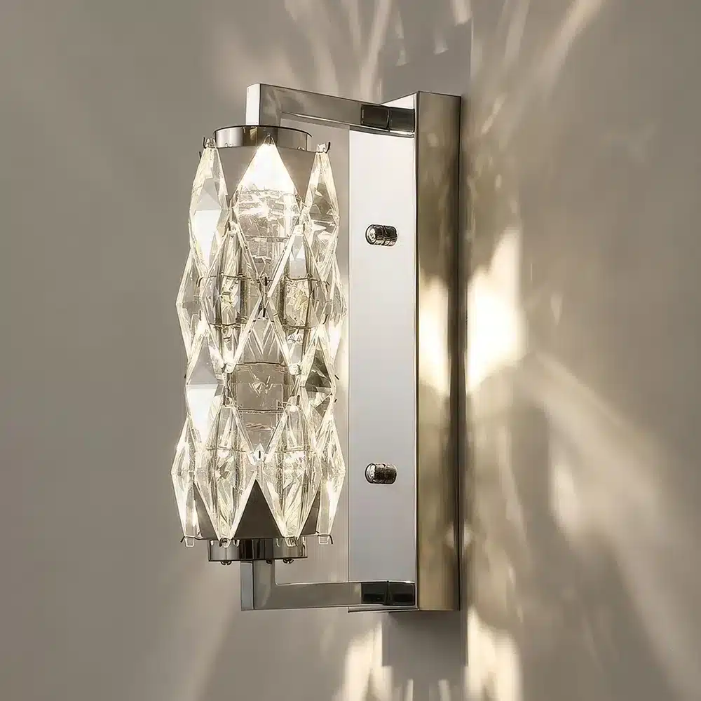 A wall-mounted light fixture with a geometric crystal design emits a warm glow, creating intricate patterns of light and shadow on the wall. The frame is metallic, complementing the modern, elegant appearance of the sconce.