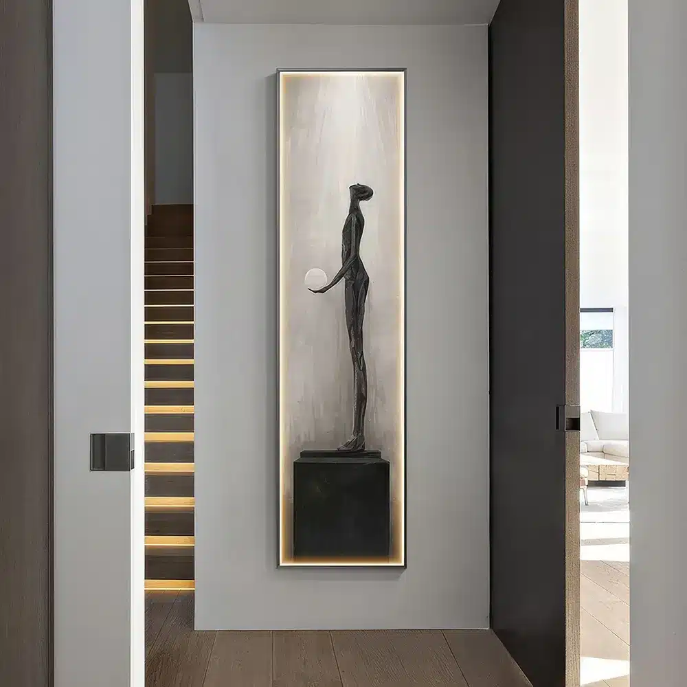 A modern hallway features an elongated, abstract sculpture of a standing figure holding a sphere. The artwork is displayed in a tall, illuminated niche, with wooden stairs on the left and a glimpse of a living area on the right.