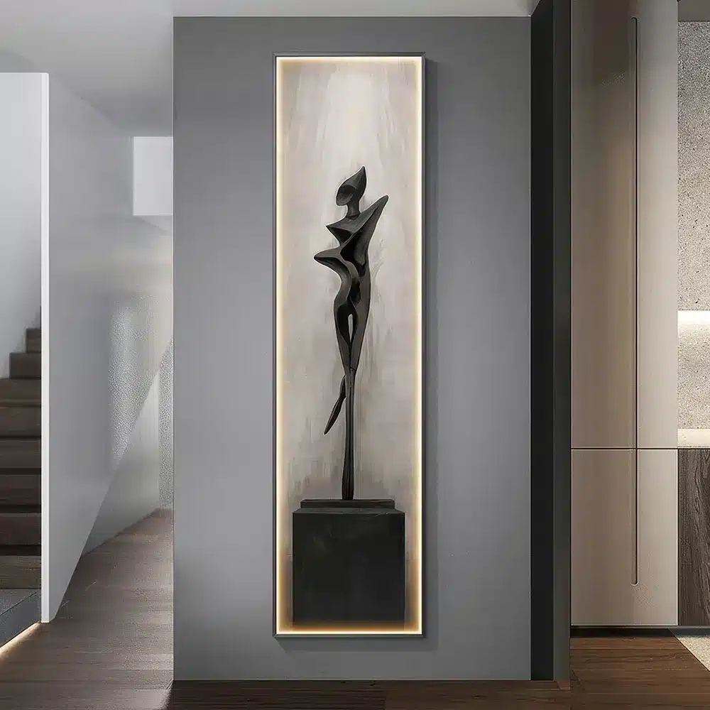 A modern interior features a tall, slim black sculpture on a rectangular pedestal. The sculpture is abstract, with a stylized human form, and is framed by soft lighting. The room has minimalist decor with stairs and a wall in the background.