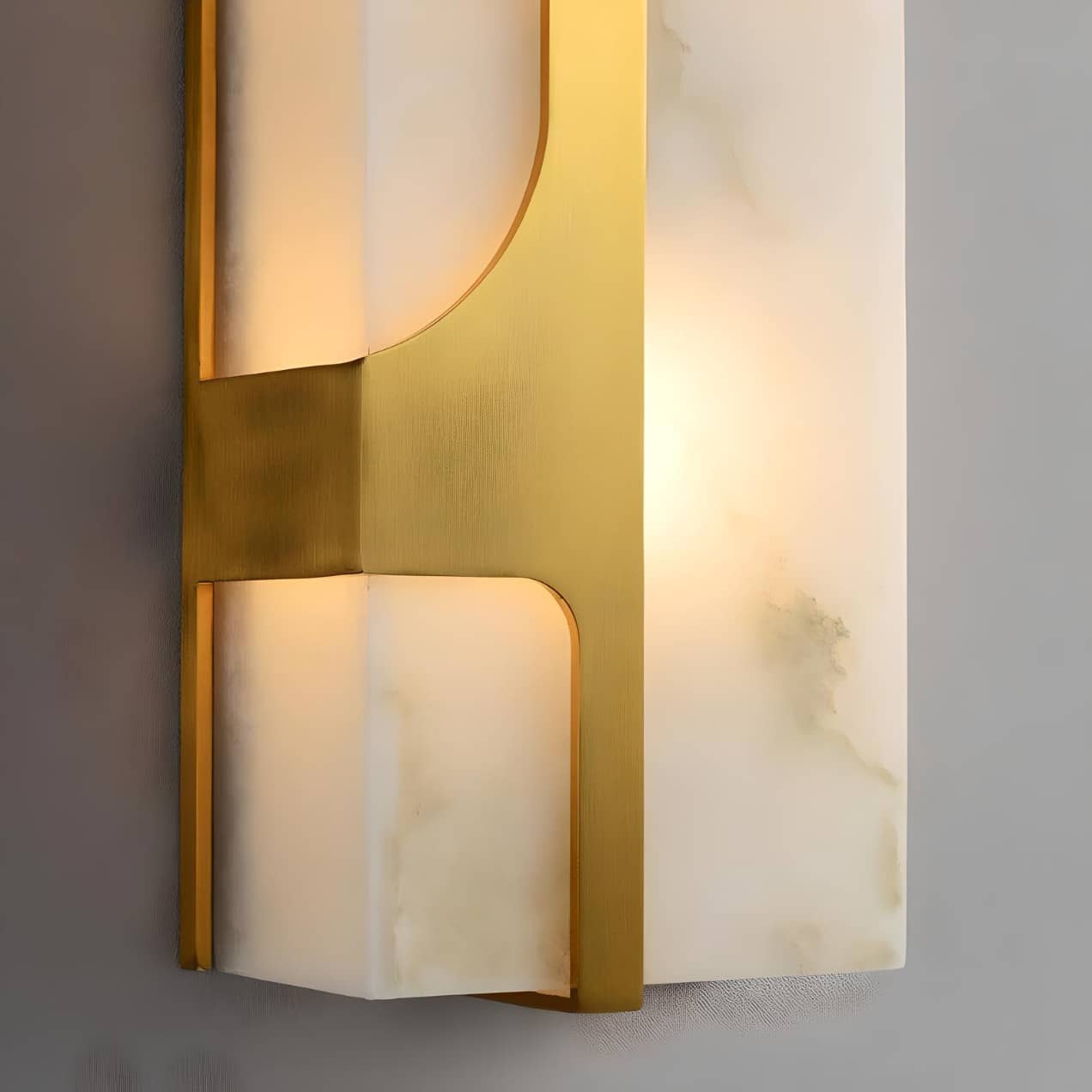 Close-up of a modern wall sconce with a rectangular white shade framed in gold metal. The light creates a warm glow against a neutral background, highlighting the elegant design and texture of the materials.