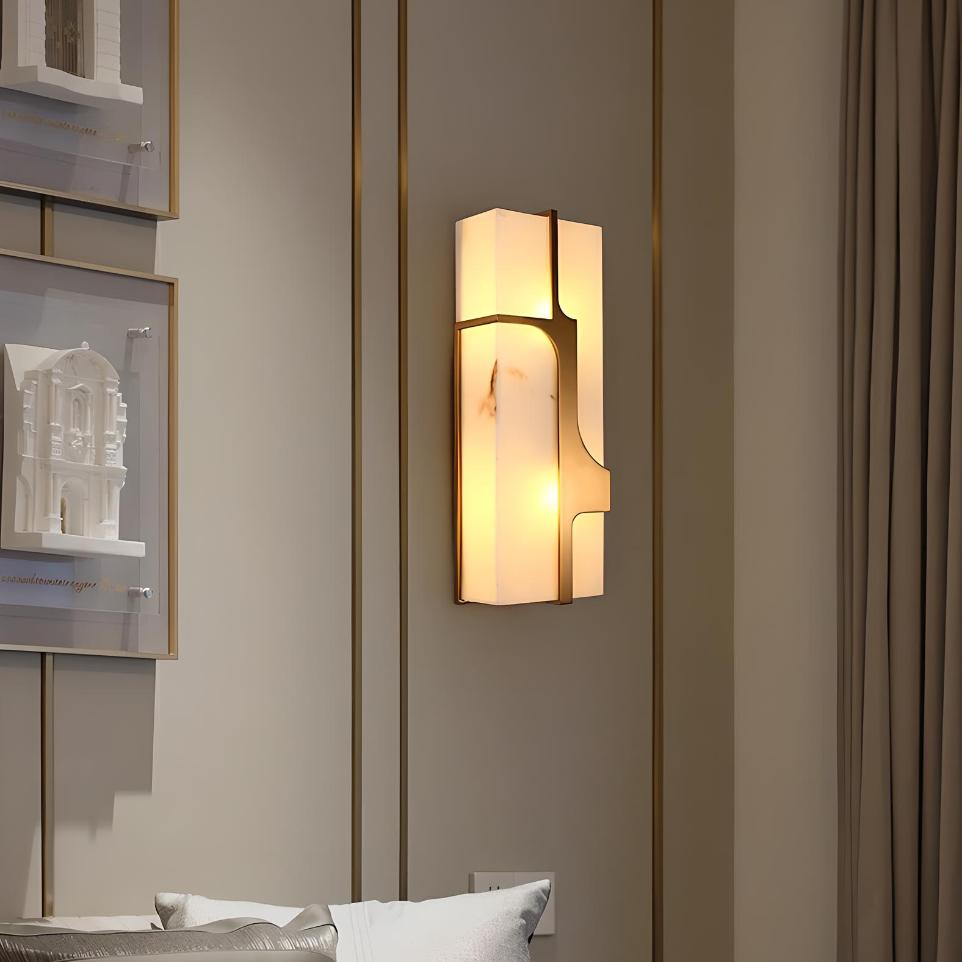 A modern wall-mounted light fixture illuminates a room with soft, warm light. The sleek design features geometric patterns and a frosted finish. The surrounding walls are neutral-toned with framed artwork, and a portion of a bed is visible.