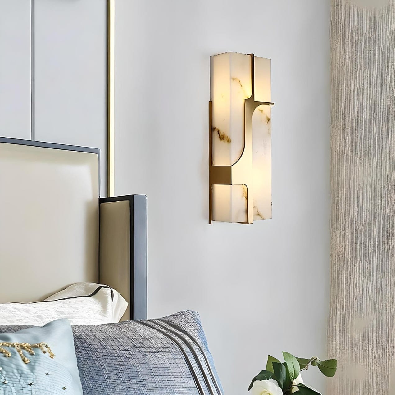 A modern bedroom features a sleek, geometric wall sconce with a gold frame and frosted glass. The corner of a bed is visible, adorned with blue and gray bedding. A potted plant is placed nearby, adding a touch of greenery.