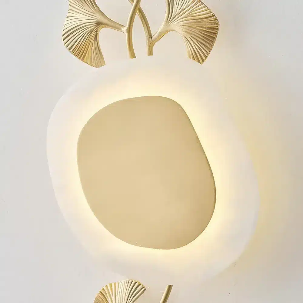 A modern wall mirror with a soft, irregular oval shape, framed by a glowing backlight. Decorative gold leaves accent the top and bottom, adding an elegant touch.