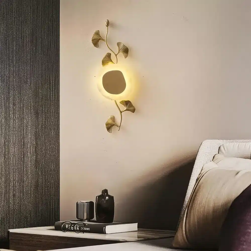 A stylish wall lamp designed like a sun with surrounding leaves casts a warm glow in a modern bedroom. Below, a table holds decorative items, including a black abstract sculpture and stacked books.