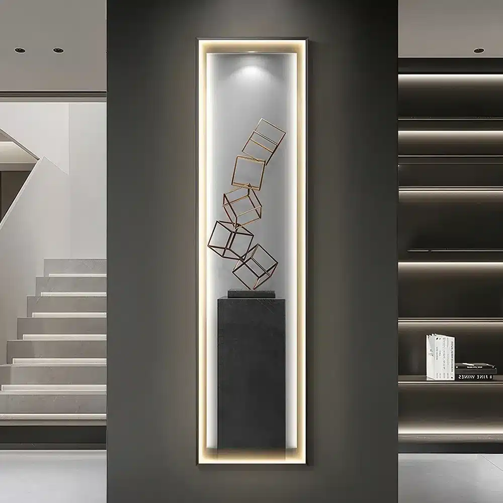 A tall, narrow wall display features a series of metal cubes balanced on top of a black pedestal, encased in a backlit frame. The setup is flanked by a staircase on the left and shelves on the right.