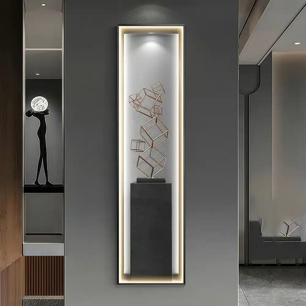 A modern hallway features a tall, narrow illuminated display case. Inside, a sculptural arrangement of interconnected metal cubes is mounted on a black pedestal. The adjacent room shows minimalistic decor with a monochrome color scheme.