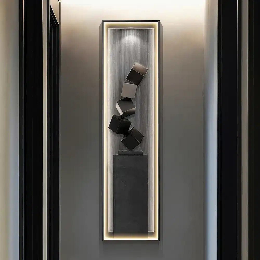 A tall, narrow sculpture featuring a series of black cubes stacked in a twisting formation, displayed within a recessed, illuminated wall niche. The surrounding walls are dark, accentuating the sculptures modern design.