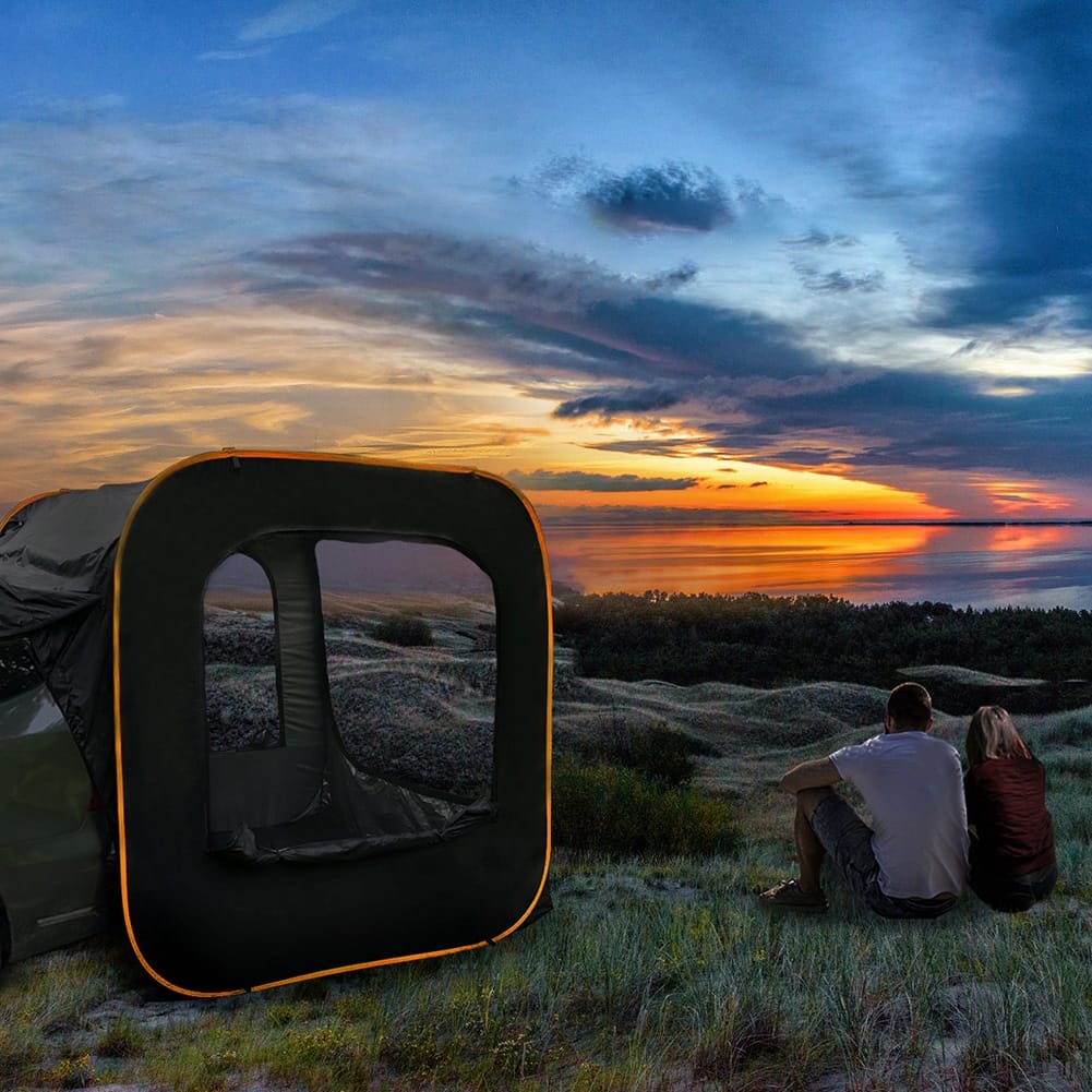 A couple sits on grass near a tent, overlooking a picturesque sunset. The sky is filled with dramatic clouds and vibrant hues of orange and blue, casting a warm glow over the landscape.