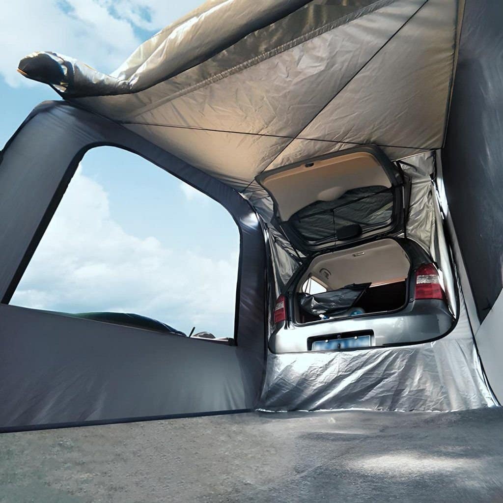 A car with its trunk open is connected to a gray tent, providing a sheltered space. The tent extends from the back of the vehicle, blending outdoor camping with car accessibility. A blue sky with scattered clouds is in the background.