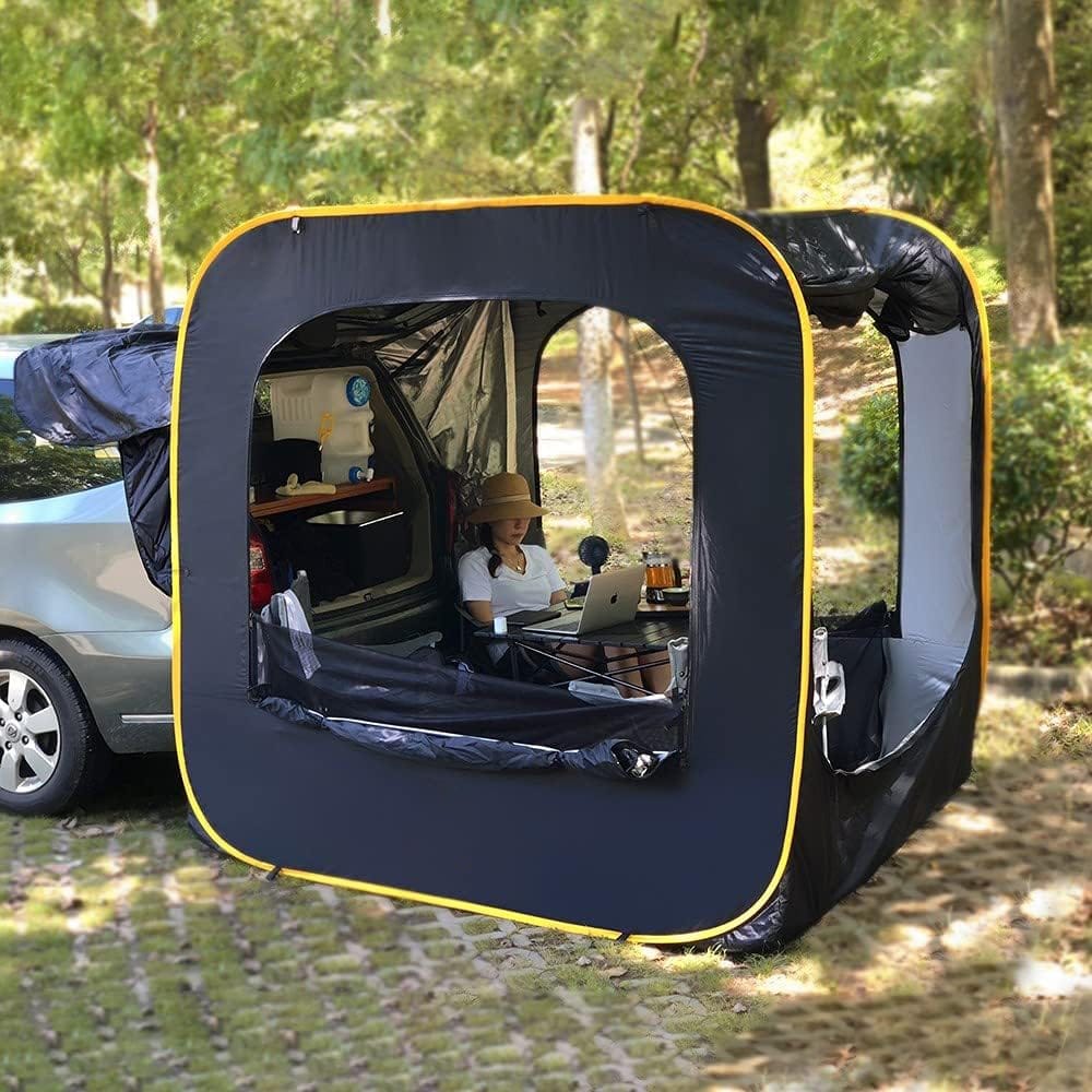 A person wearing a hat is sitting on a folding chair inside a large tent attached to a car. The tent has open windows, and the person is using a laptop. The car is parked on grassy pavement, and trees are visible in the background.