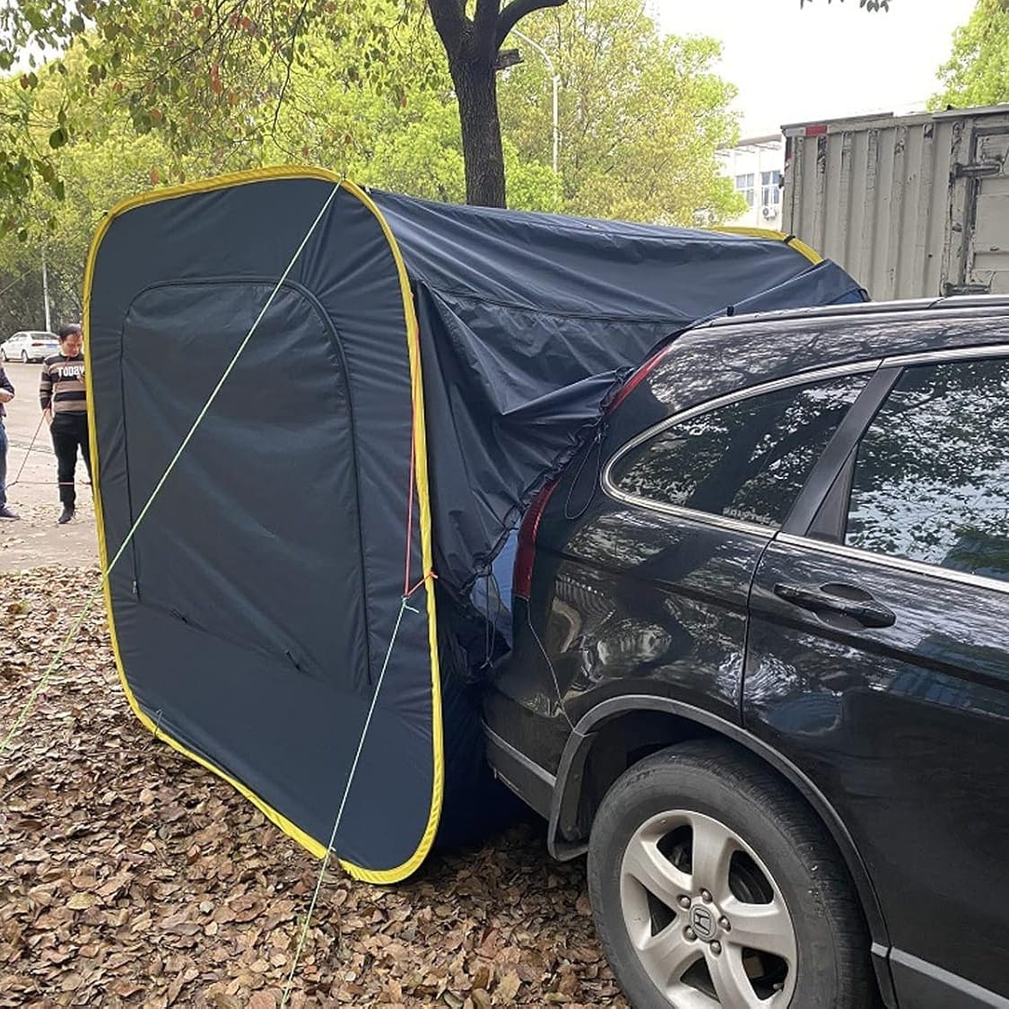 A dark blue tent is attached to the rear of a parked black SUV, extending from the cars open hatch. The tent is secured with ropes and stakes on a leaf-covered ground. Trees line the background, with a person walking in the distance.