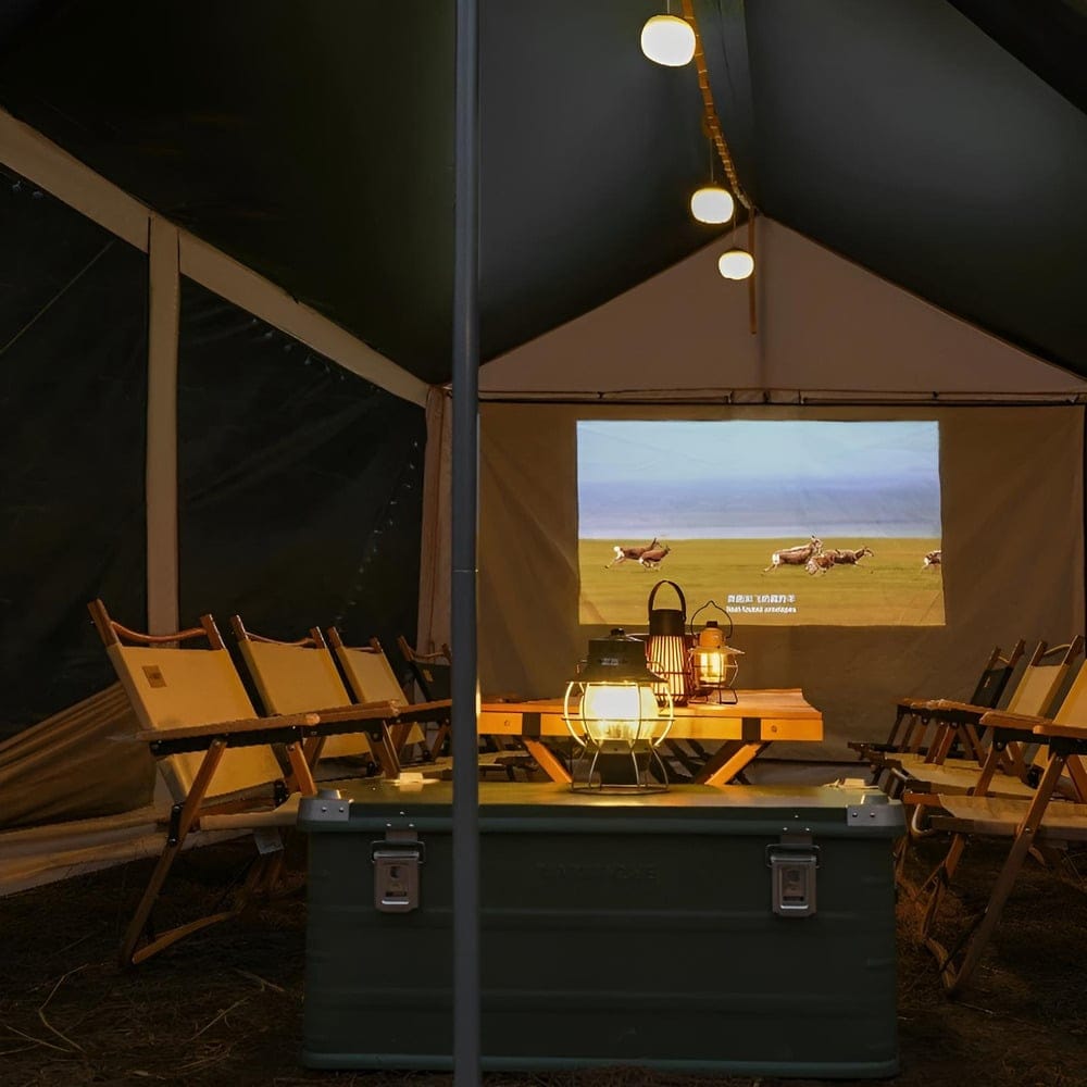 Interior of a tent set up for a movie night, featuring a screen showing a landscape scene, folding chairs, a wooden table with a lantern, and string lights overhead, creating a cozy atmosphere.