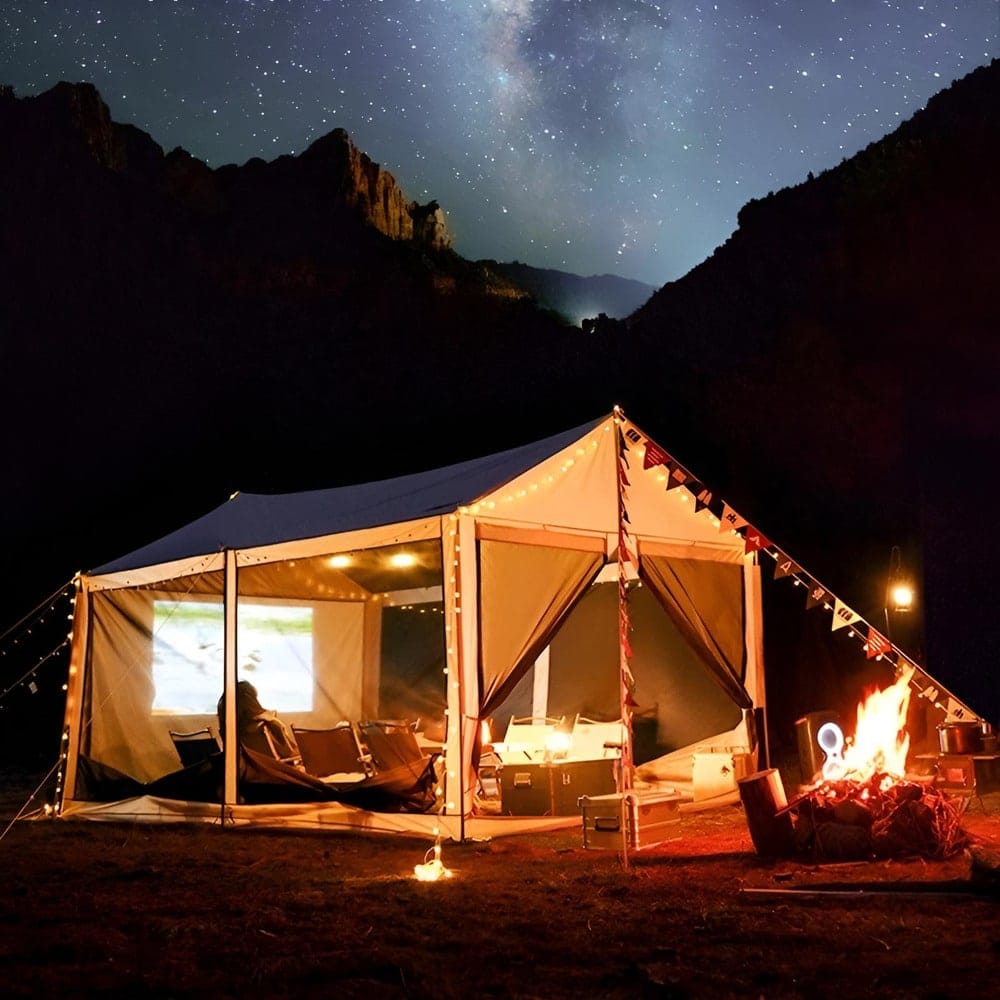 A large canvas tent stands illuminated with string lights at night, set in a mountainous landscape. The tent interior shows comfortable seating, while a bright campfire burns outside. A starry sky and the Milky Way create a serene backdrop.