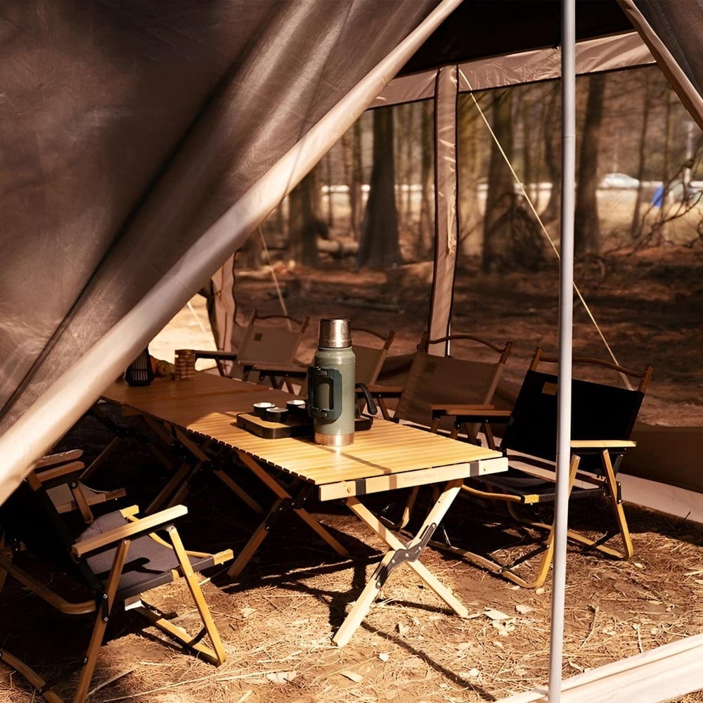 Inside view of a tent with a camping table and folding chairs. A thermos and mug sit on the table. Sunlight filters through the trees outside, casting shadows on the ground. The scene conveys a cozy outdoor atmosphere.