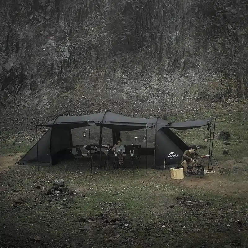 A large, dark camping tent is set up in a rocky outdoor area. People are sitting under the canopy, surrounded by camping gear. The atmosphere is overcast and rugged, suggesting a remote, wilderness location.