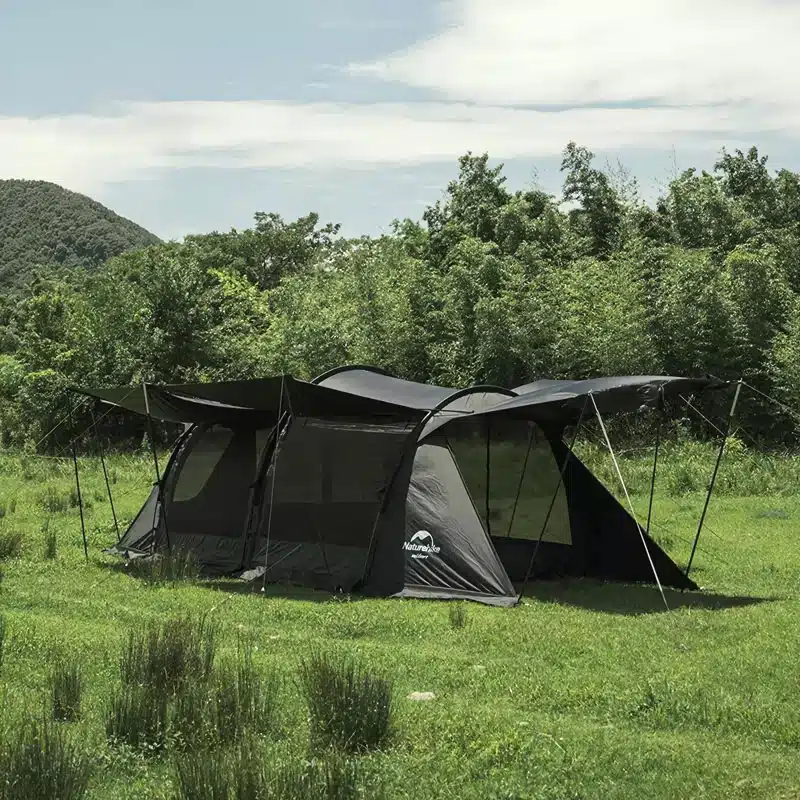 A large, dark-colored camping tent is set up on a grassy field surrounded by lush green trees. The sky is partly cloudy, and a small hill is visible in the background.