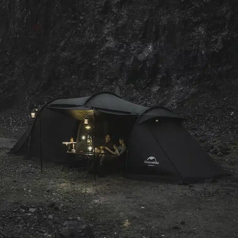 A nighttime scene showing a dark outdoor tent illuminated by several lanterns. Two people are sitting inside the tent, surrounded by rocky terrain. The atmosphere is cozy and calm.