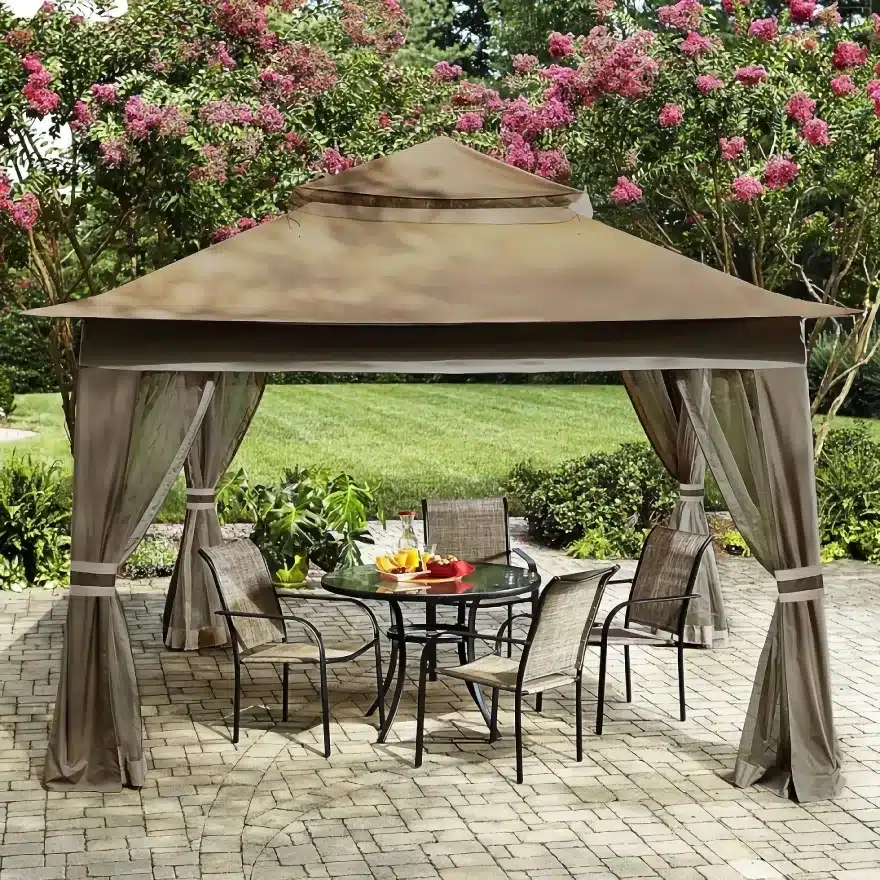 A patio scene featuring a tan gazebo with sheer curtains tied to its posts, covering a round glass table with a fruit platter and four chairs. The setting is on a stone patio surrounded by lush greenery and blooming pink trees.