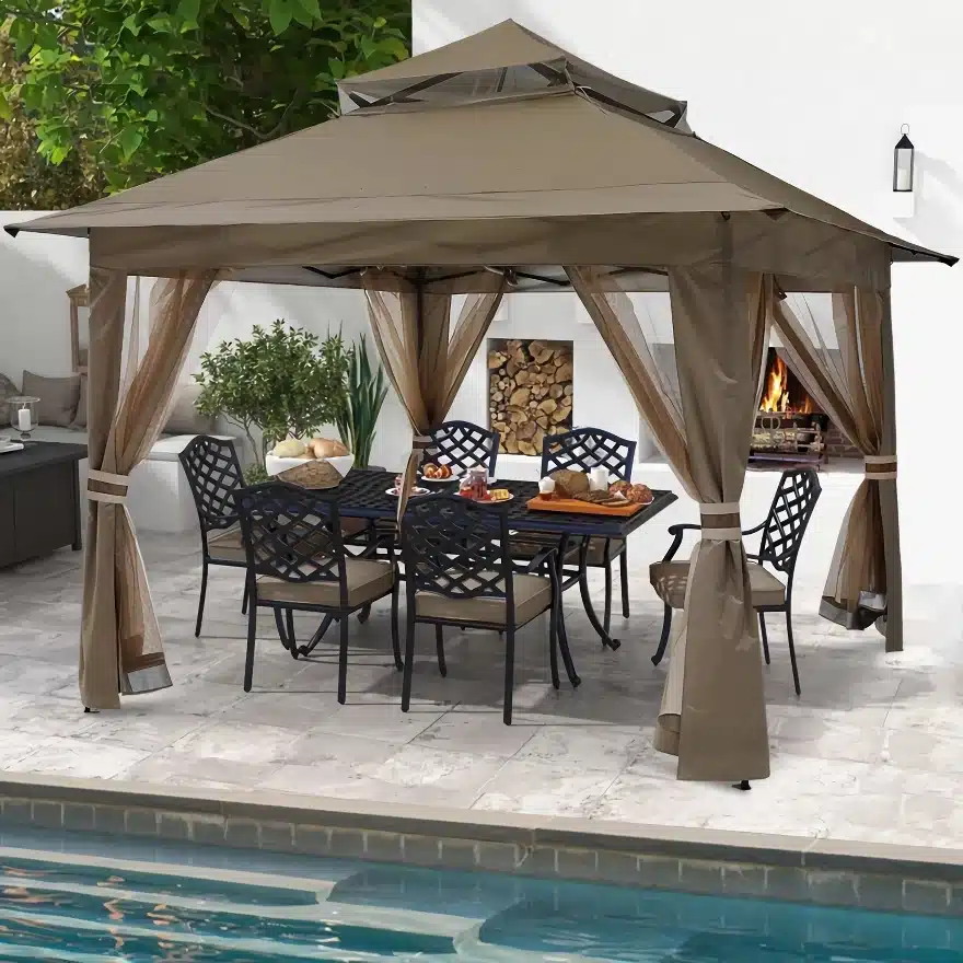 A brown canopy tent covers a patio dining area beside a pool. The table is set with plates of food, and cushioned chairs surround it. A fireplace and stacked firewood are visible against a white wall with greenery in the background.