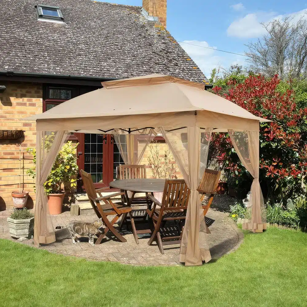 A beige canopy tent covers a wooden dining set with six chairs on a patio in a garden. A cat walks nearby. The background features a house with a brick exterior and green shrubs under a clear blue sky.