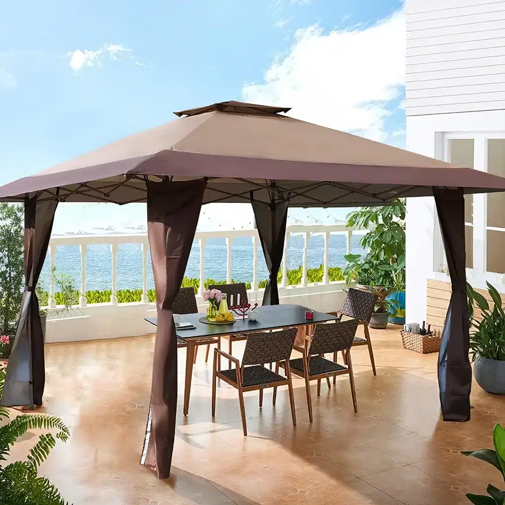 A patio with a brown canopy gazebo covering a dining table and six chairs. The area overlooks a body of water, surrounded by a railing. Theres some potted greenery, and the sky is blue with a few clouds.