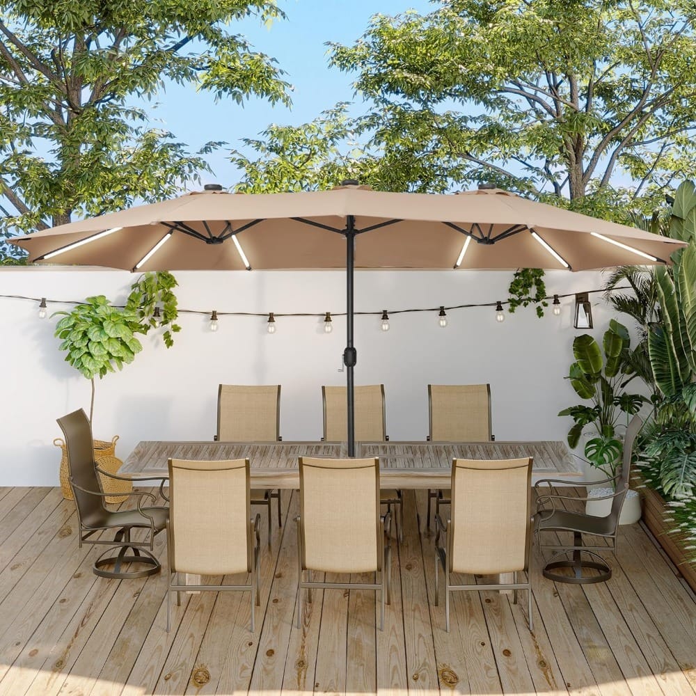 A patio setup with a large tan umbrella covering a wooden table and eight beige chairs. String lights and potted plants hang on a white wall in the background. The area is surrounded by trees, creating a serene outdoor setting.