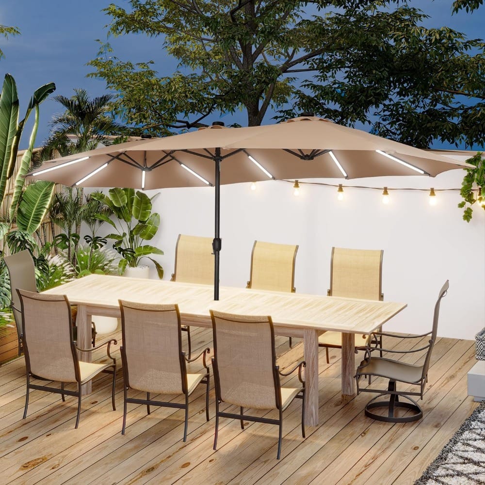 A wooden deck features a long dining table with eight beige chairs. An overhead umbrella with string lights provides shade. The area is decorated with tropical plants and additional string lights, creating a cozy outdoor setting.